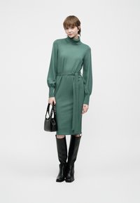 Grønt, strikket kjole med lange ermer, høy krage og belte i livet. Et par svarte knelang støvler og en liten svart håndveske.