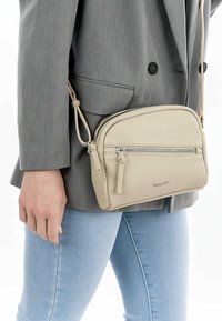 Borsa a tracolla in pelle beige con chiusura a zip e dettagli con cerniera, caratterizzata da una texture morbida e una tracolla regolabile, indossata sopra un blazer grigio.