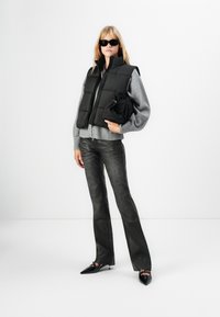 Vrouw met zwarte zonnebril, zwarte puffer vest over een grijze trui, zwarte leren broek, houdt een zwart opgevouwen jack vast, staande op een witte achtergrond.