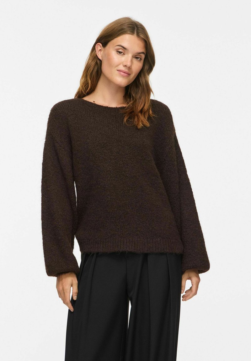 Femme portant un pull en laine pelucheux marron foncé à manches longues et un pantalon noir à jambes larges, debout devant un fond clair.