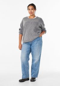 Harmaa neulepaita, jossa on rento muotoilu, pyöreä pääntie ja lasketut olkapäät. Se on yhdistetty vaaleansinisiin korkeavyötäröisiin farkkuihin ja mustiin nilkkureihin.