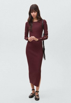 Rochie burgundy din material cu dungi, cu mâneci lungi, fanta pe lateral și detalii cu nasturi pe mâneci, asortată cu pantofi negri și o geantă mică de umăr.