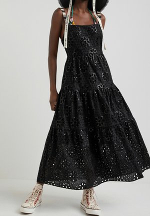 Kobieta w czarnej, warstwowej sukience z ażurowymi kwiatowymi wycięciami, w białych sneakersach typu high-top z czerwonymi akcentami, stojąca na jednolitym tle.