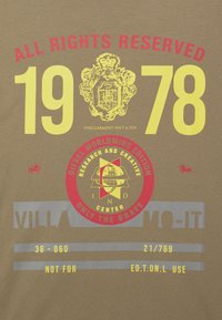 Olivově zelené bavlněné tričko s grafickými potisky v červené, žluté a šedé barvě, včetně roku "1978" a různých log a textů v tučných písmech.