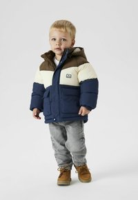 Giacca puffy blu navy e crema con dettagli marroni e cappuccio, dotata di tasche laterali. Indossata con jeans grigi chiari e stivali marroni.