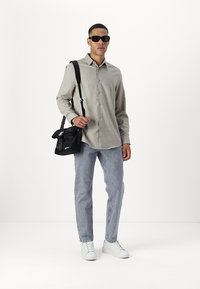 Camisa de botones beige claro, jeans azul claro, zapatillas blancas y un bolso cruzado negro. La persona lleva gafas de sol oscuras.