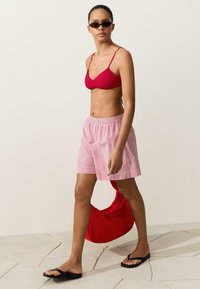 Femme marchant portant un haut de bikini rouge, un short rayé rose et blanc, des tongs noirs, des lunettes de soleil et portant un sac rouge.