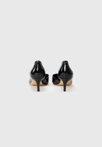 Ein Paar schwarze Lackleder-Mid-Heel-Pumps von hinten auf einer weißen Fläche mit beigen Sohlen und schmalen Absätzen.