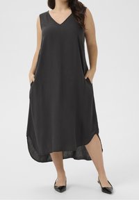 Femme portant une robe midi sans manches noire avec un décolleté en V et des poches latérales, associée à des chaussures noires à bout pointu sur un fond uni.