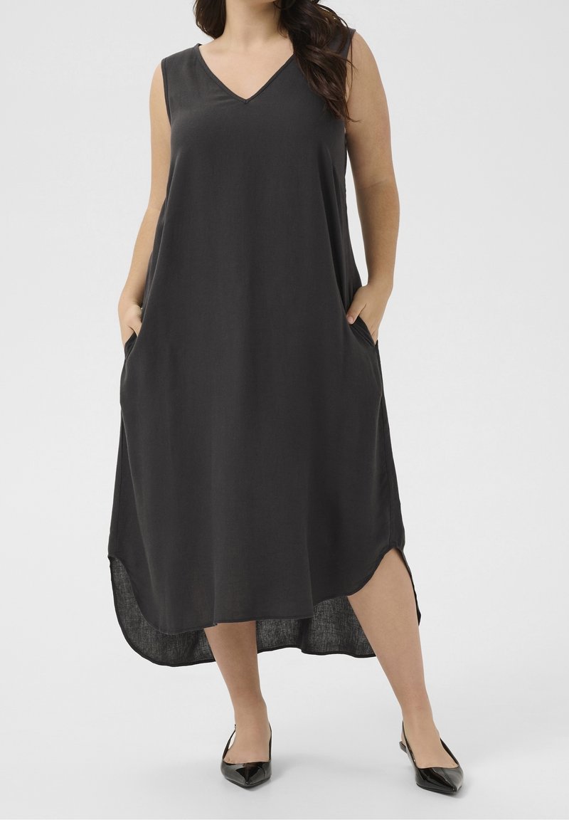 Femme portant une robe midi sans manches noire avec un décolleté en V et des poches latérales, associée à des chaussures noires à bout pointu sur un fond uni.