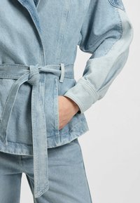 Light blue denim jacket with wide sleeves, belted waist, and a pocket. The fabric shows a smooth texture and a slightly faded finish.
