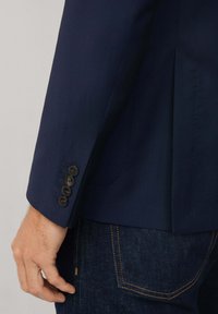 Granatowa marynarka o gładkiej fakturze, z pojedynczym rozcięciem i pięcioma ciemnymi guzikami na mankietach. Zestawiona z ciemnymi dżinsami.