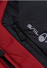 Röd och svart isolerad jacka med en dragkedja, rektangulärt sömnads mönster, broderad logotyp och slät textur.