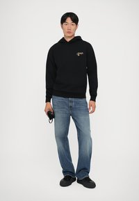 Hoodie en coton noir avec logo, poche avant et poignets côtelés, associé à un jean en denim bleu clair et des chaussures noires, tenant un sac dans la main droite.