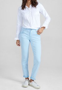 Pantalon bleu clair coupe slim avec une texture lisse, doté de poches avant et de revers roulés, associé à une chemise blanche à manches longues boutonnée.