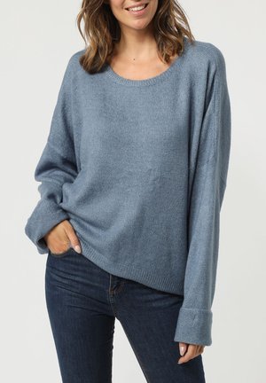 Jersey de punto azul claro con un corte holgado, cuello redondo y mangas largas con puños anchos. Combinado con jeans oscuros.