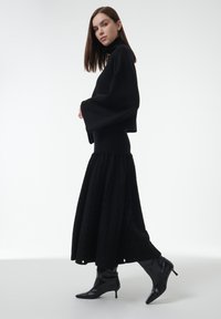 Vrouw in een zwarte turtlenecktrui, lange textuurrok en zwarte hoge laarzen die tegen een effen witte achtergrond loopt.