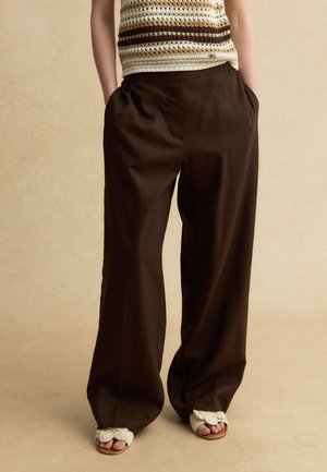 Persona che indossa pantaloni marrone scuro a zampa larga, top senza maniche a righe lavorato all'uncinetto e sandali aperti con cinturini crema testurizzati. Mani in tasca.