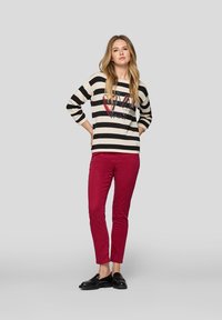 Gestreifter Pullover in Schwarz und Creme mit einem grafischen Herzensdesign, kombiniert mit figurbetonten burgunderroten Hosen und schwarzen Loafers, an einem Modell.