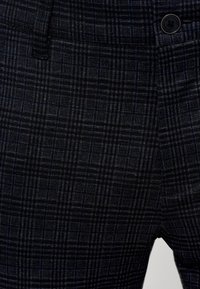 Pantalon gris foncé avec un motif à carreaux subtil, doté d’un bouton à l’avant et d’un petit détail sur la poche avant.