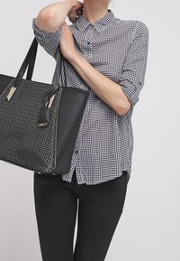 Blusa a quadri bianchi e neri con colletto, abbinata a una borsa tote nera testurizzata con un motivo geometrico e dettagli dorati.
