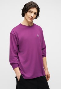 Νεαρός με μεσαίου μήκους σκούρα μαλλιά, φορώντας μπλούζα με μώβ μακριά μανίκια και μικρό τριγωνικό λογότυπο στο στήθος, με τα χέρια στις τσέπες.