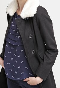 Manteau noir avec un col en fausse fourrure blanche, présentant un design croisé, de gros boutons et une coupe cintrée, porté sur un chemisier à volants.