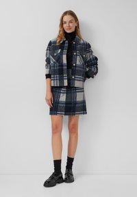 Karierte Wolljacke in Marineblau und Grau mit Knopfverschluss und zwei Brusttaschen, kombiniert mit einem passenden karierten Minirock und schwarzen Loafers.