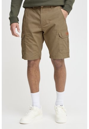 Homme portant un short cargo marron, des baskets blanches et des chaussettes blanches montantes, debout avec une main dans la poche sur un fond uni.