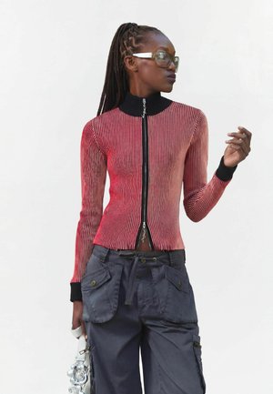 Frau mit geflochtenem Haar, trägt große Brille, rot-schwarz gestreiftes Zip-up-Oberteil, dunkle Cargo-Hose, hält eine silberne Tasche mit Blumendetails