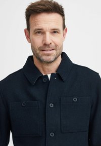 Dark navy button-up jacket with a textured fabric. Features a collar and two chest pockets. The design is simple and utilitarian.