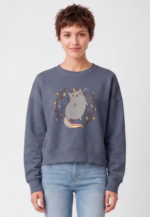 Jeune femme aux cheveux courts portant un jean bleu et un sweat-shirt bleu avec un chat licorne dessiné avec une queue arc-en-ciel et des branches fleuries.