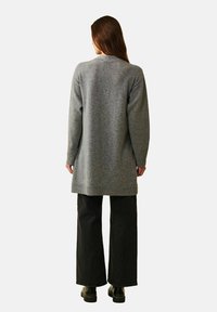 Lång grå stickad cardigan med ribbad fåll och muddar, bärs över svarta vida byxor. Texturerat tyg, minimalistisk design, bakifrån.