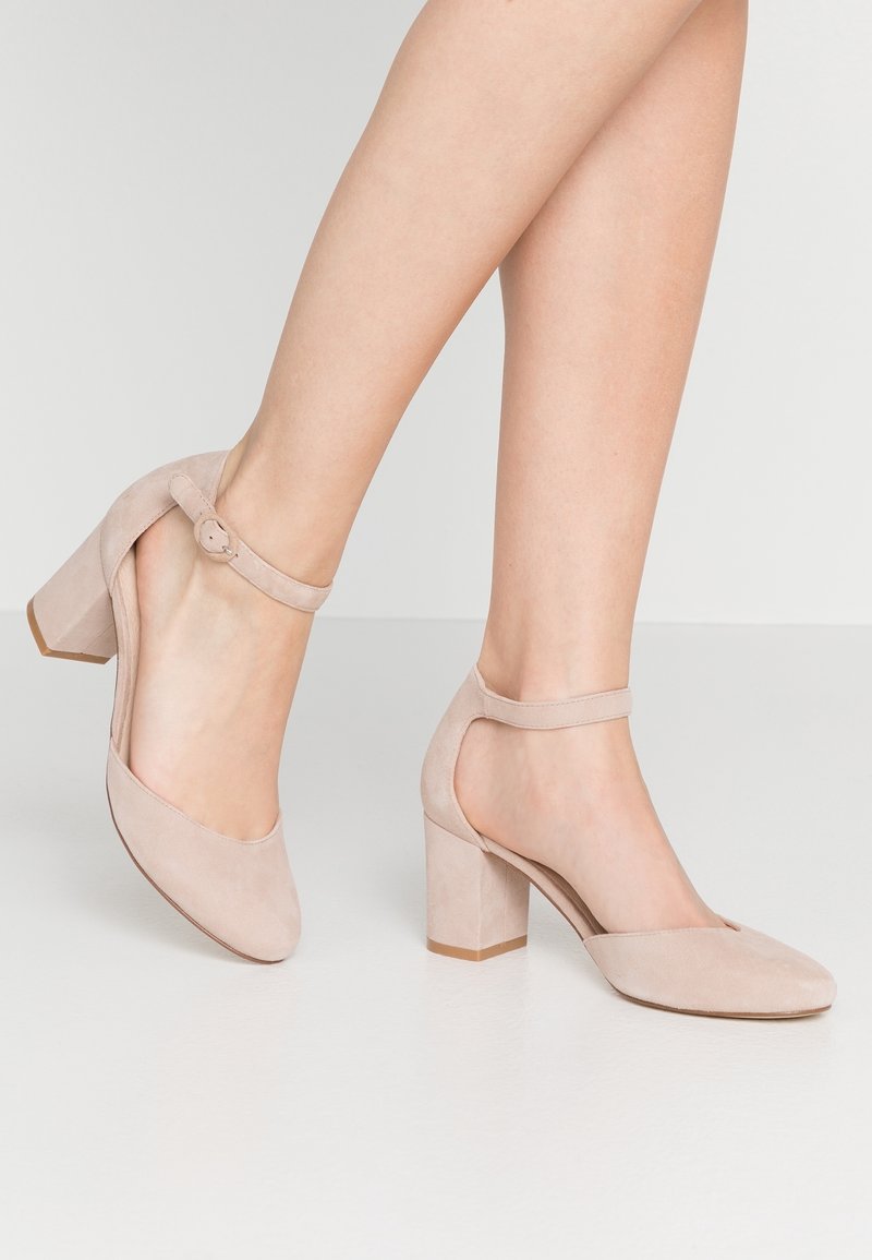 Jambes portant des escarpins à talons blocs en daim beige avec des brides à la cheville sur un fond neutre.