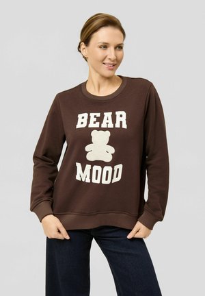 Vrouw draagt een bruine trui met een witte teddybeer-afbeelding en tekst "BEAR MOOD", staand tegen een effen lichte achtergrond.