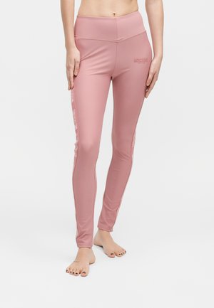 Nő, magas derekú rózsaszín leggingset visel „Moschino Active” logóval, mezítláb áll egy egyszínű fehér háttér előtt.