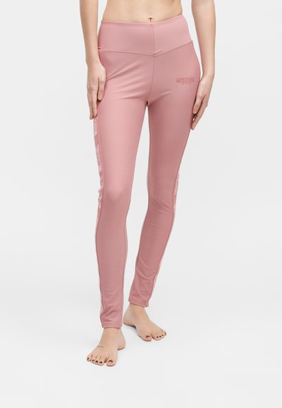 Femme portant un legging rose taille haute avec le logo « Moschino Active », debout pieds nus sur un fond blanc uni.