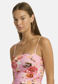 Une jeune femme aux longs cheveux porte une robe florale rose à fines bretelles, regardant l'objectif contre un fond blanc uni.