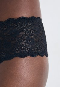 Fekete csipkeből készült alsónemű, fodros felső szélekkel, virágmintákkal és puha tapintással, amely kiemeli a részletes tervezést.