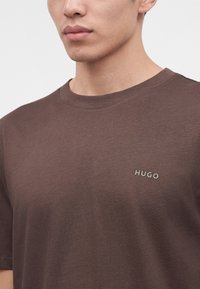 Homme portant un t-shirt marron uni avec un petit logo "HUGO" sur le côté gauche de la poitrine, montré des épaules à la bouche.
