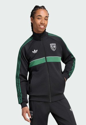 Junger Mann mit geflochtenem Haar, der eine schwarze Adidas-Trainingsjacke und eine Hose mit grünen Streifen und Racing Club de Lens-Logos trägt, lächelt mit den Händen in den Taschen.