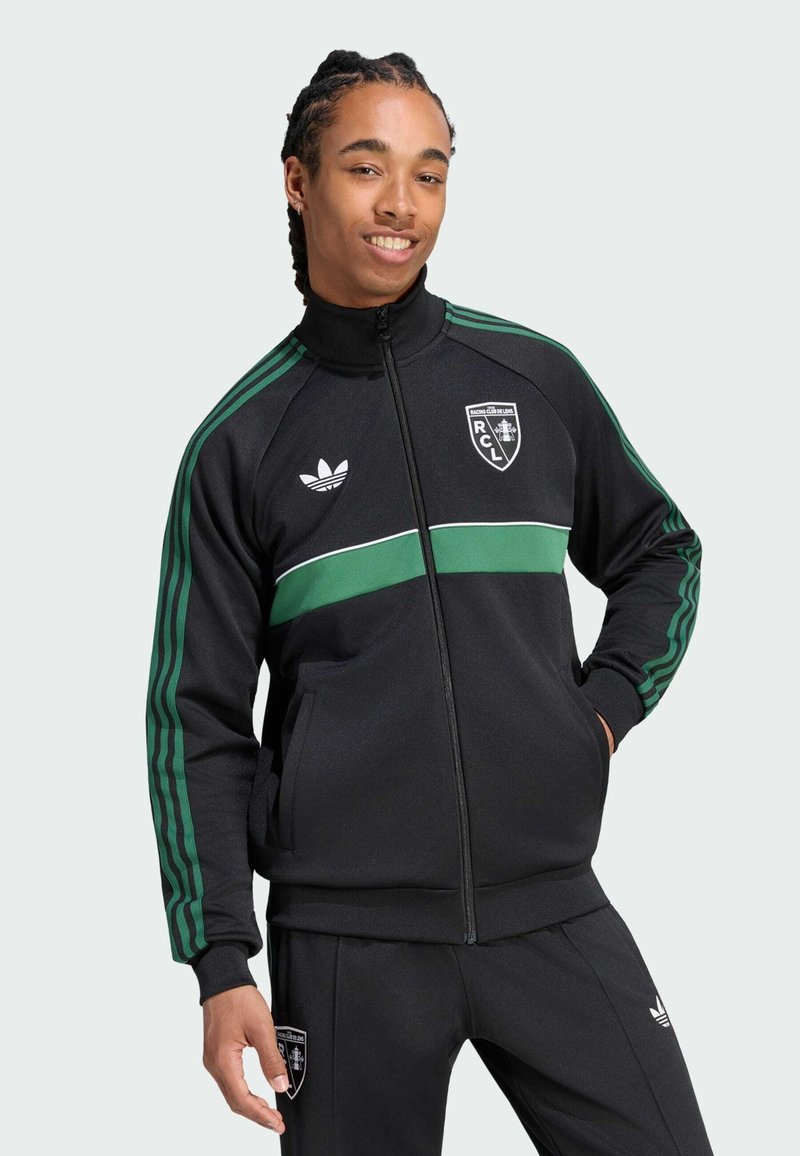 Jeune homme aux cheveux tressés portant une veste et un pantalon de survêtement noirs Adidas avec des bandes vertes et des logos du Racing Club de Lens, souriant les mains dans les poches.