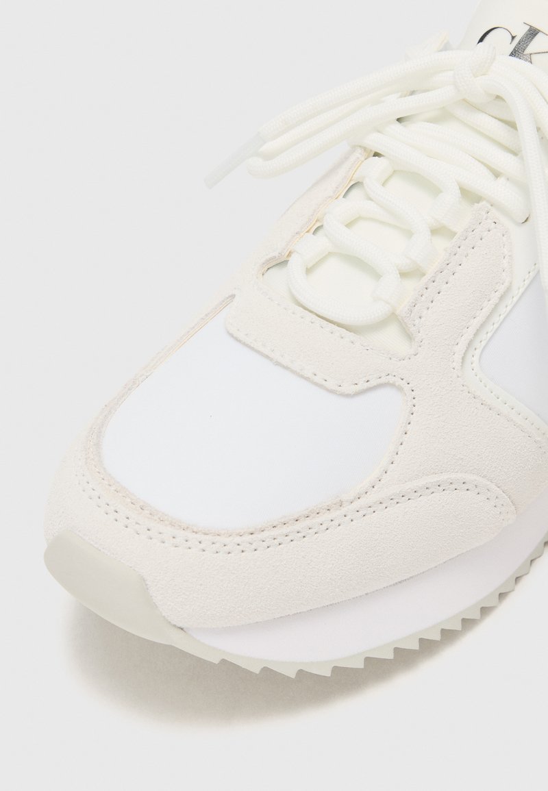 Zapatilla blanca con una mezcla de materiales lisos y texturizados, que presenta una puntera de ante, diseño de cordones y una suela de goma estriada.