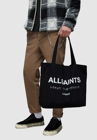 Black tote bag with "ALLSAINTS" logo, made of cotton, featuring two sturdy handles. Worn with brown pants and black leopard-print shoes.