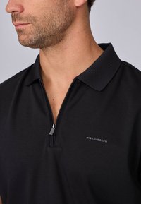 Polo noir avec fermeture éclair, col côtelé et un petit logo blanc. Le tissu semble lisse avec un léger éclat.