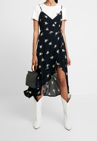 Vestido envolvente negro con estampado floral y tirantes finos, con un dobladillo asimétrico. Llevado sobre una camiseta blanca y combinado con botas de vaquero blancas y una bolsa pequeña.