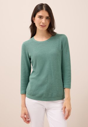 Haut vert à manches longues avec un motif tricoté texturé, encolure ronde et ourlet légèrement arrondi. Porté avec un pantalon blanc.