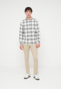 Chemise boutonnée blanche à motif à carreaux noirs, manches longues et col ; associée à un pantalon beige slim et des baskets noires et blanches.