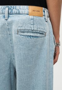 Πίσω όψη ανοιχτό μπλε τζιν με μία τσέπη με λεπτομέρεια, ταμπά ετικέτα της μάρκας "ONLY & SONS" και το χέρι ενός ατόμου που φοράει ασημένια βραχιόλια με αλυσίδα.