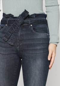 Jeans a vita alta di colore blu scuro con una cintura in tessuto annodata a fiocco, indossati con un top a maniche lunghe rigato di colore verde chiaro.